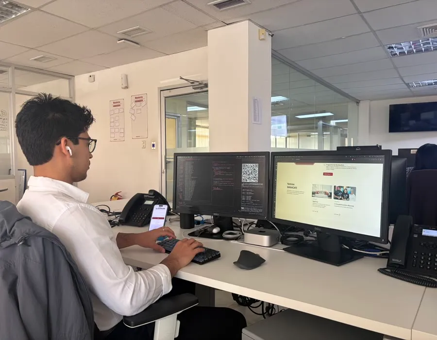 Jeral trabajando en la oficina frente a dos monitores, con código y un sitio web en pantalla