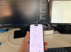 Mano sosteniendo un teléfono con una app de pagos hecha en React Native, y el código detrás en los monitores